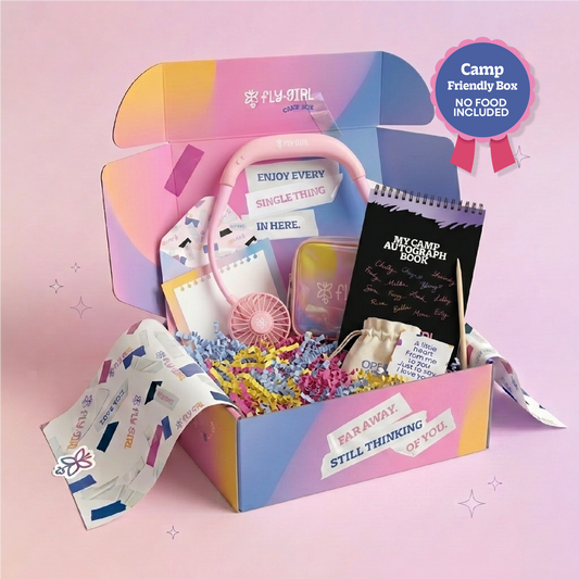 A colorful open gift box labeled 'Fly Girl' filled with assorted cute items including a pink fan, notebook, pouch, and small accessories, set against a soft pink background with a 'Camp Friendly Box No Food Included' badge.