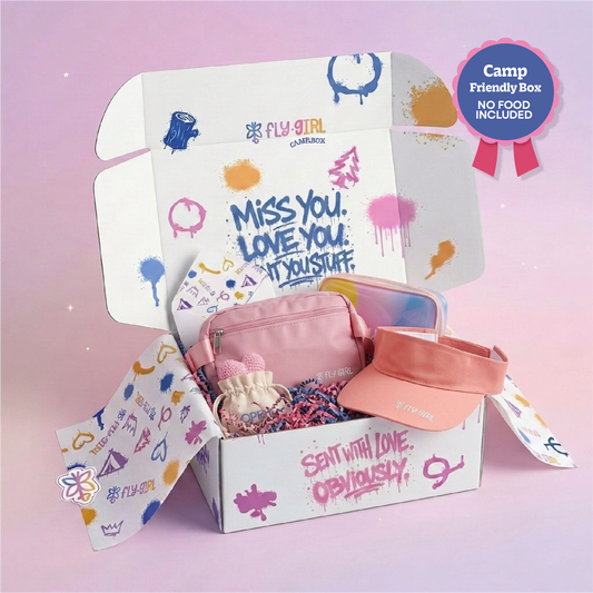 An open Fly Girl Campfire themed cardboard gift box filled with pink accessories, including a pink pouch, a peach visor, and a small plush toy, set against a soft pink background with a 'Camp Friendly Box' badge.