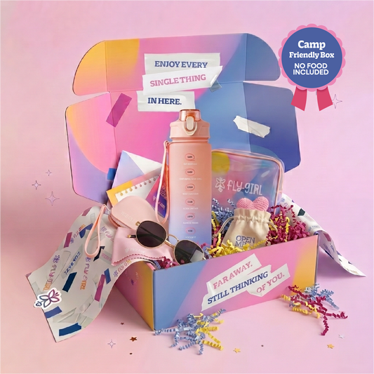 A colorful open gift box filled with travel-themed items on a pink background. Visible contents include a tall pink water bottle, a clear zip pouch labeled FLY GIRL, a small beige pouch, sunglasses, and paper accessories.