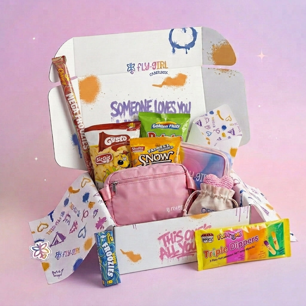A curated snack and accessory gift box set from Fly Girl Cosmetics, featuring a pink zip pouch, a bag of Skittles, a bag of Now!, a bag of Oreo, a pack of Warheads Triple Dippers, and a Warheads double pack, all arranged inside a white cardboard box with colorful graphics against a soft pink background.