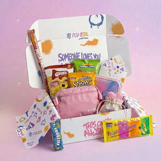 A curated snack and accessory gift box set from Fly Girl Cosmetics, featuring a pink zip pouch, a bag of Skittles, a bag of Now!, a bag of Oreo, a pack of Warheads Triple Dippers, and a Warheads double pack, all arranged inside a white cardboard box with colorful graphics against a soft pink background.
