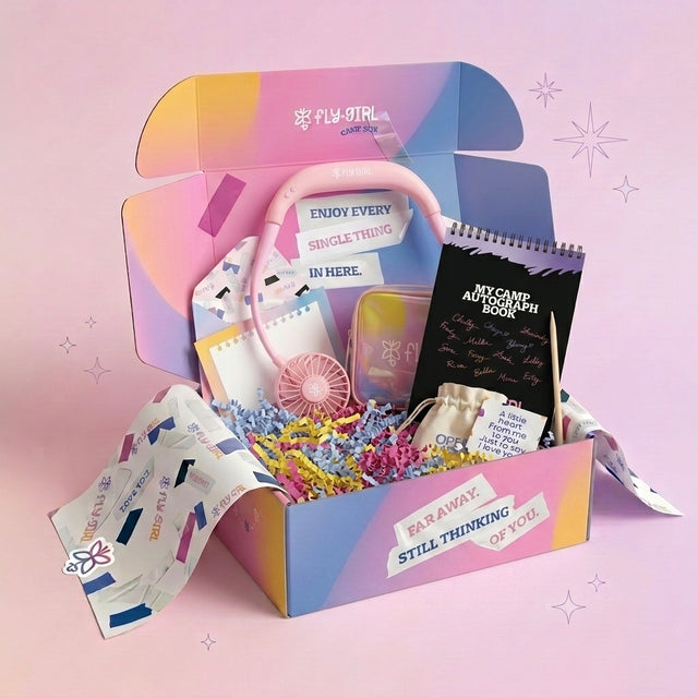 A colorful gift box set from Fly Girl on a pink background. The open box contains a pink headphone, a small pink fan, a black spiral notebook, a small pouch, and other pastel accessories arranged in crinkle paper.
