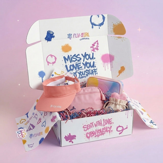 A Fly Girl Care Package gift box shown open, filled with a peach visor, pink pouches, and colorful accessories, set against a soft pink background with decorative sparkles.