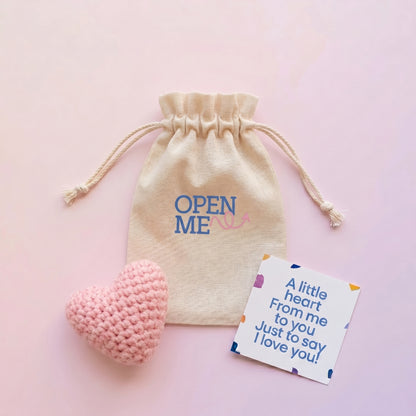 Beige drawstring bag with 'OPEN ME' text, pink heart-shaped object, and card on a pink background